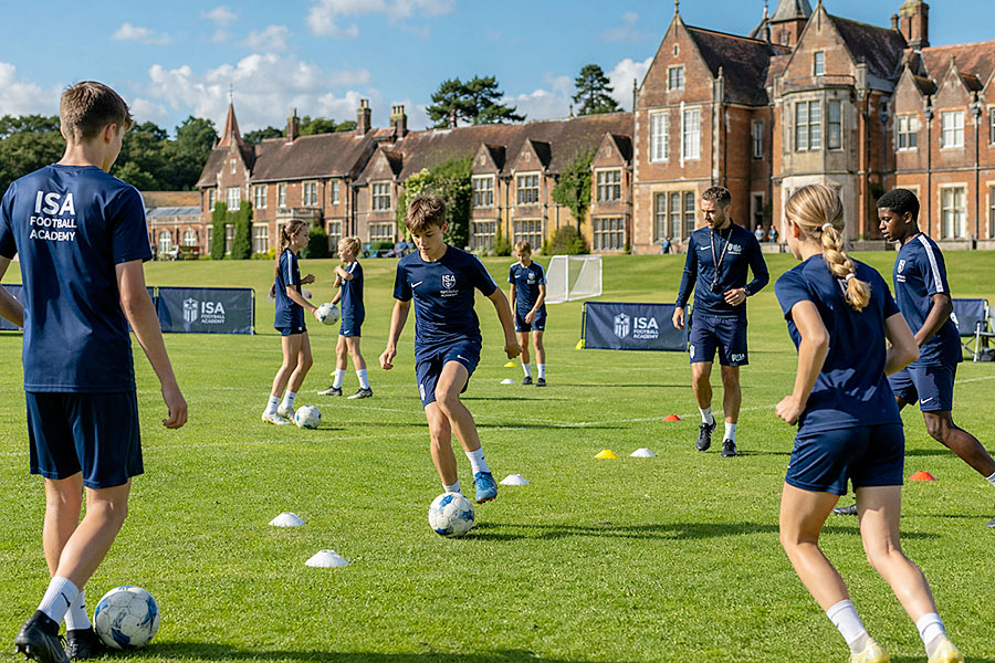 Un gruppo di adolescenti in uniforme sportiva blu, parte di un'Accademia di calcio, si esercita su un campo erboso con coni e palloni all'esterno di un grande edificio storico in una giornata di sole.