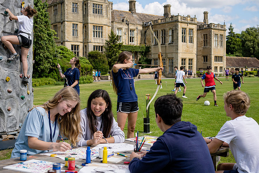 Gli adolescenti si divertono con varie attività all'aperto in un vivace campo multiattività su un prato verde davanti a un grande edificio storico; alcuni dipingono su un tavolo, uno si arrampica su una parete di roccia, un altro pratica il tiro con l'arco, mentre altri giocano a calcio sullo sfondo.