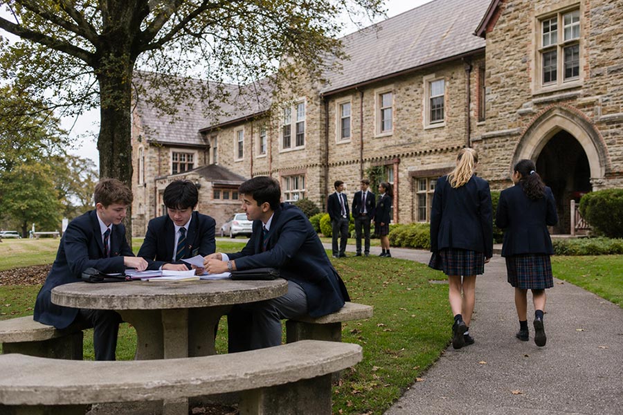 Quattro studenti in uniforme siedono a un tavolo rotondo di pietra per studiare all'esterno di un grande edificio scolastico in mattoni, una scena ideale per chi vuole studiare un anno in Nuova Zelanda, con altri che camminano lungo il sentiero sullo sfondo.