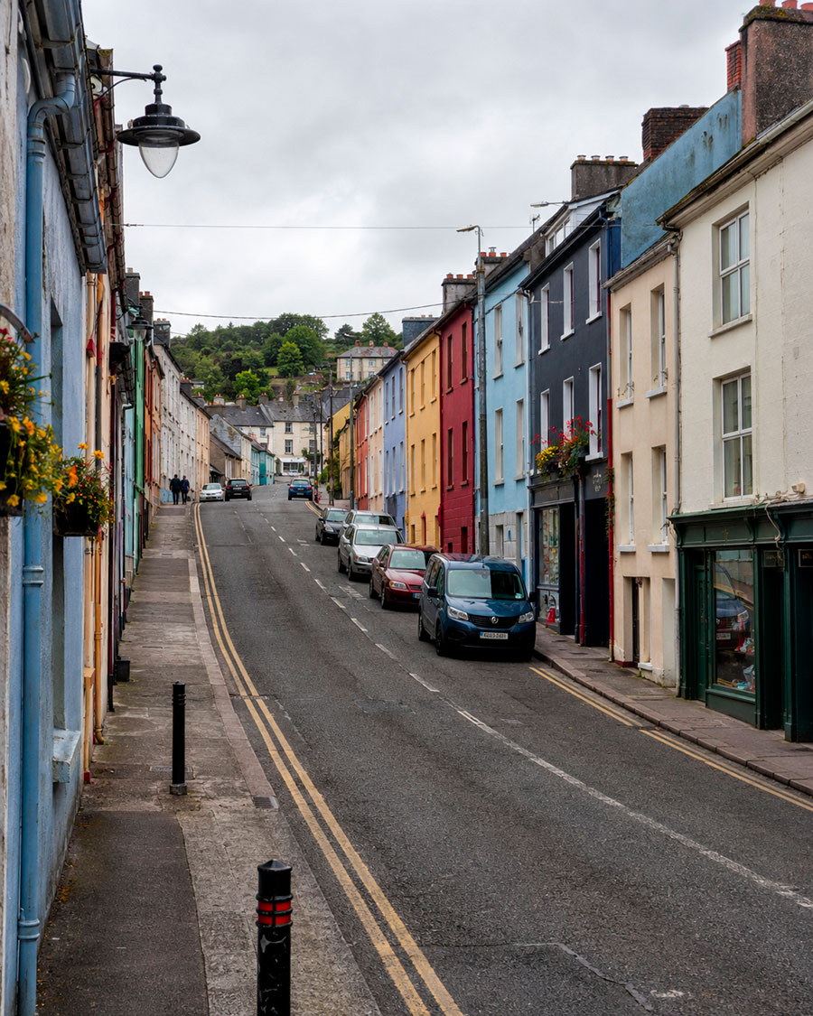 Una strada stretta in Irlanda, fiancheggiata da edifici colorati a due piani e auto parcheggiate, cattura il fascino che gli studenti potrebbero sperimentare durante il loro anno scolastico irlandese. Cesti di fiori appesi decorano alcune facciate mentre la strada sale sotto un cielo nuvoloso.