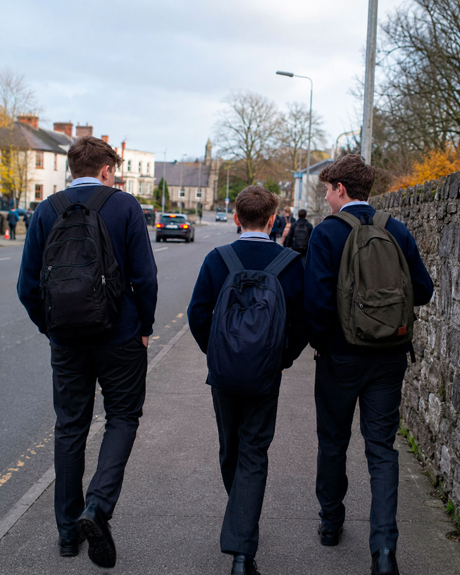 Tre studenti in uniforme scolastica e zaino in spalla camminano insieme su un marciapiede accanto a un muro di pietra, con alberi ed edifici visibili lungo la strada in una giornata nuvolosa - una scena quotidiana durante un anno scolastico irlandese.