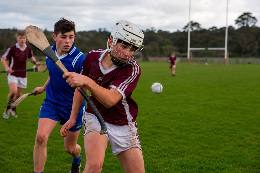Due ragazzi giocano a hurling su un campo erboso: uno in uniforme maroon e casco bianco fa oscillare la sua hurley, mentre un altro in blu corre dietro. La scena cattura l'eccitazione dello sport durante un anno scolastico irlandese, con le porte sullo sfondo.