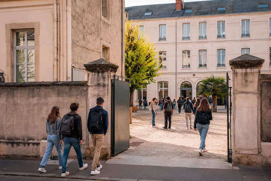 Studenti con lo zaino in spalla attraversano un cancello aperto per entrare nel cortile di una scuola, circondata da edifici chiari: una scena tipica di chi sta vivendo un anno scolastico in Francia, magari durante il suo quarto anno francese.