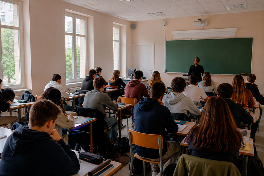 Un'aula in Francia con gli studenti seduti ai banchi di fronte all'insegnante, davanti a una grande lavagna verde, perfetta per chi sta vivendo un anno scolastico in Francia, con alte finestre che riempiono la stanza di luce naturale.