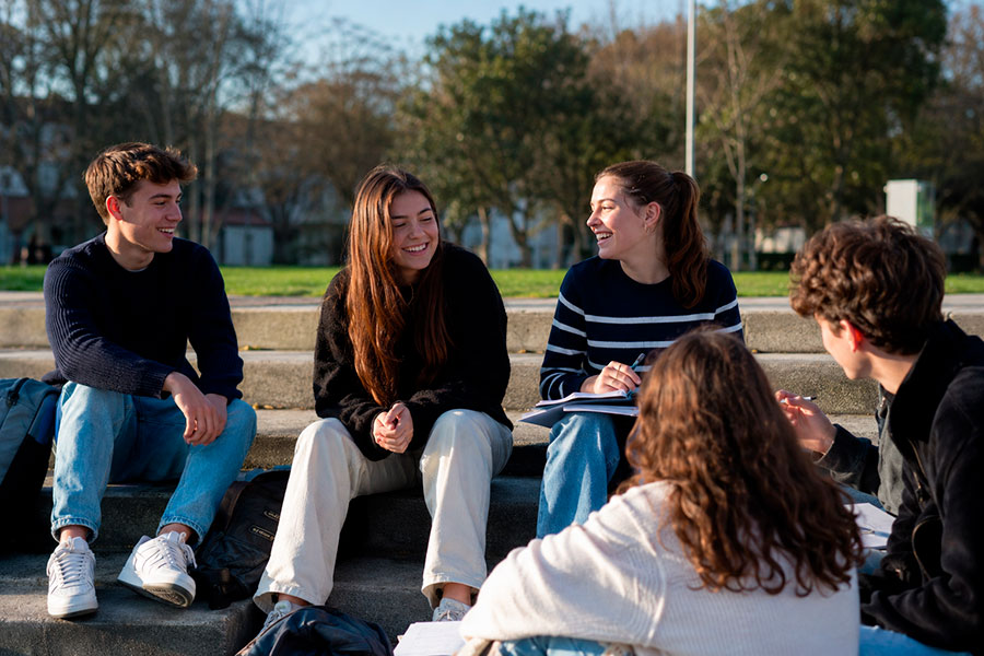 Un gruppo di cinque giovani adulti siede sui gradini all'aperto, parlando e ridendo insieme. Sembrano rilassati e amichevoli, con quaderni e borse nelle vicinanze, godendosi una giornata di sole in un parco durante il loro anno scolastico in Irlanda.