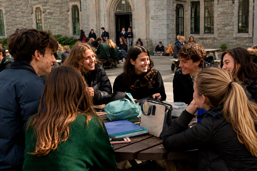 Un gruppo di studenti siede intorno a un tavolo da picnic in legno all'esterno di un edificio in pietra, sorridendo e parlando del loro anno scolastico irlandese. Sul tavolo ci sono quaderni, borse e cartelle. Sullo sfondo del cortile ci sono altre persone.