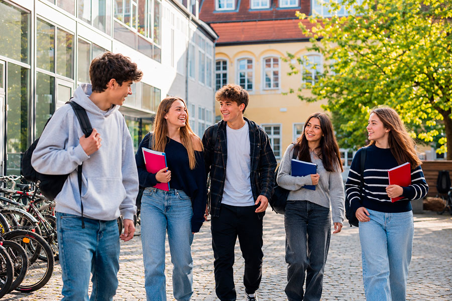 Cinque studenti sorridenti camminano insieme all'aperto in un campus scolastico, chiacchierando delle loro esperienze nell'anno scolastico tedesco. Portano con sé dei quaderni e si godono la giornata di sole, con biciclette ed edifici sullo sfondo.