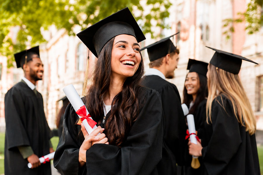 Un gruppo di giovani laureati in abito da sera si trova all'aperto, sorridente e con i diplomi in mano, con un edificio del campus verdeggiante sullo sfondo. Una donna in primo piano sembra allegra ed eccitata dopo un orientamento universitario di successo.