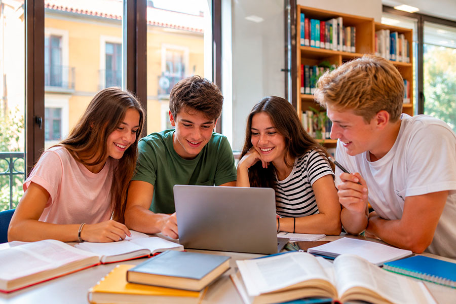 Quattro giovani adulti sorridenti siedono a un tavolo in una biblioteca, studiando insieme mentre discutono del loro anno all'estero Spagna. Guardano un computer portatile e hanno davanti a sé libri e quaderni aperti, con la luce del sole che filtra da grandi finestre alle loro spalle.