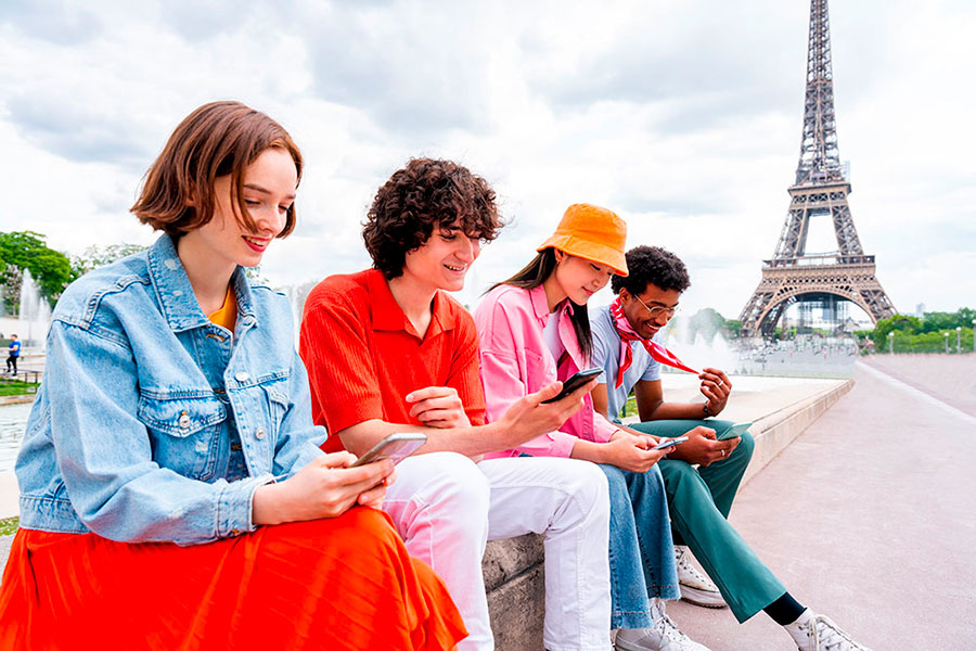 Quattro giovani siedono su un cornicione vicino alla Torre Eiffel a Parigi, sorridendo ai loro smartphone. È una giornata luminosa, perfetta per catturare i ricordi durante la loro avventura Anno scolastico Francia.