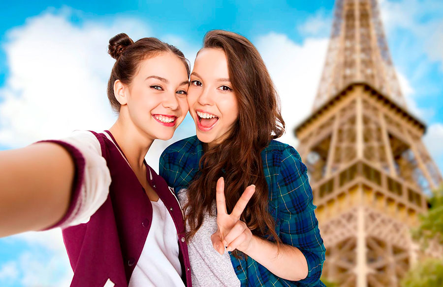 Due giovani donne sorridono e si scattano un selfie davanti alla Torre Eiffel, una con la macchina fotografica in mano e l'altra che fa un segno di pace, catturando i ricordi del loro anno scolastico in Francia. Il cielo è azzurro con alcune nuvole sullo sfondo.