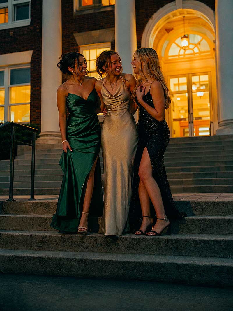 Tre giovani donne in eleganti abiti da sera stanno insieme su una scalinata all'aperto, di notte, sorridendo e parlando, con l'ingresso illuminato di un edificio con colonne e finestre ad arco sullo sfondo.