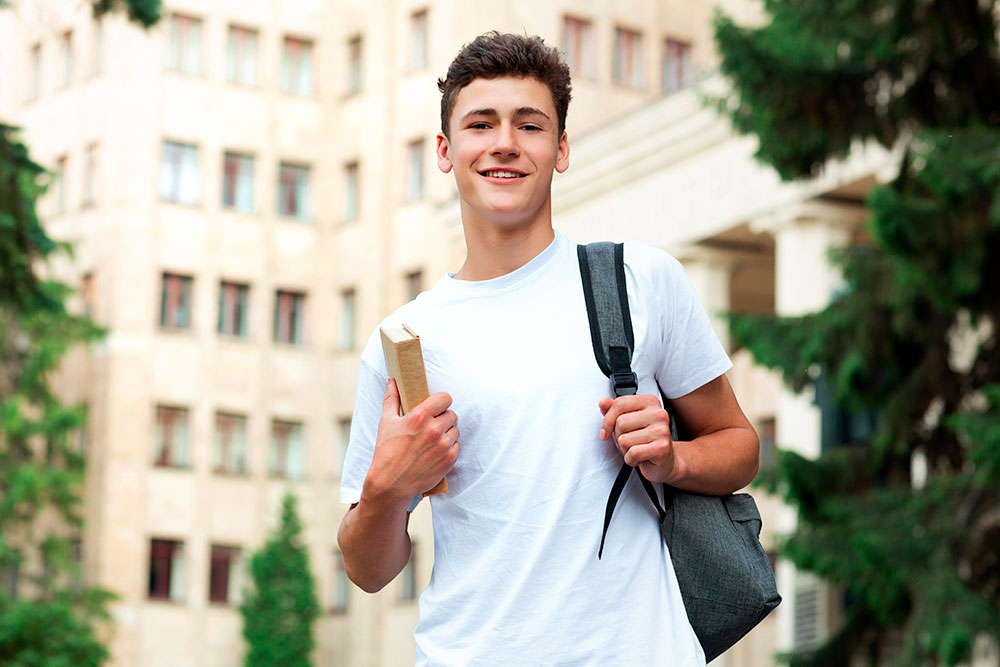 Un ragazzo sorridente con una maglietta bianca tiene in mano un libro e indossa uno zaino; è in piedi all'aperto, con degli alberi e un grande edificio alle sue spalle, e sembra pronto per l'esame, con la fisarmonica in mano.
