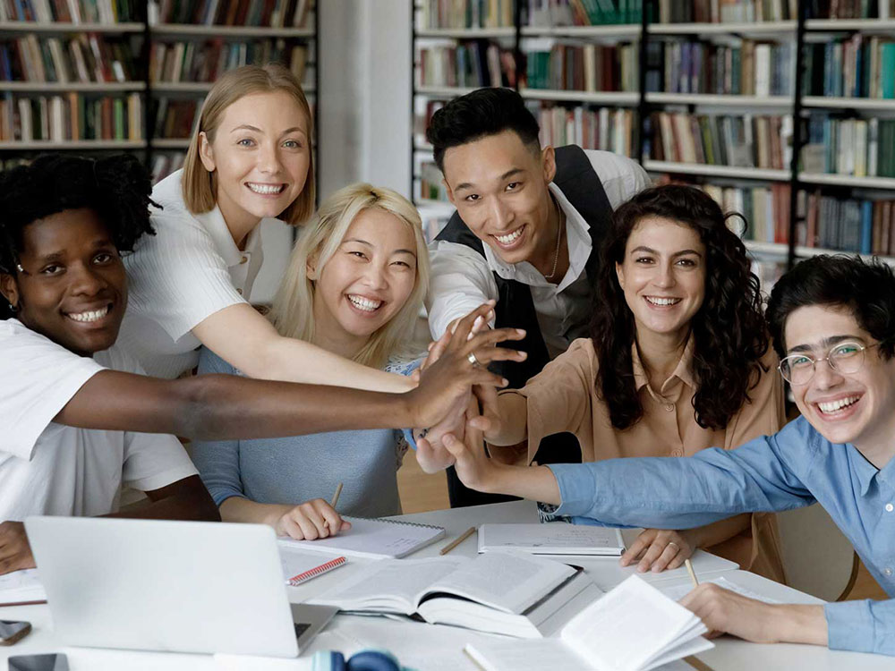 Un gruppo eterogeneo di sei studenti sorridenti siede a un tavolo in una biblioteca, allungando le mani su libri e quaderni aperti, mostrando lavoro di squadra e unità.