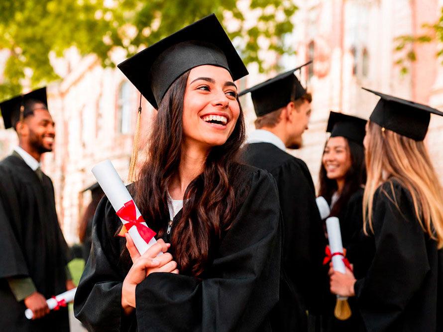 Un gruppo di laureati che indossano cappellini e camice nere si trovano insieme all'aperto. Una giovane donna in primo piano sorride luminosa, tenendo in mano un diploma legato con un nastro rosso. Sullo sfondo sono visibili alberi e un edificio.