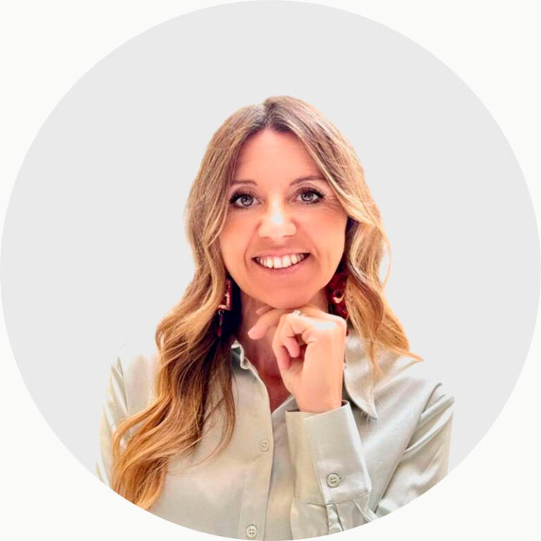 Donna sorridente con lunghi capelli biondi ondulati che indossa una camicia chiara con bottoni, in posa con la mano appoggiata sotto il mento e una fisarmonica posta nelle vicinanze, su uno sfondo semplice di colore grigio chiaro.
