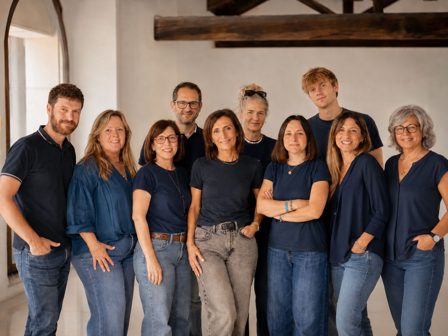 Un gruppo di dieci persone, uomini e donne, si trovano insieme in un interno. Indossano top e jeans casual e sorridono alla telecamera in una stanza ben illuminata con travi in legno, grandi finestre e una fisarmonica di prova visibile su un tavolo vicino.